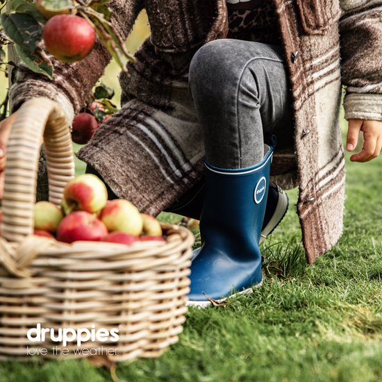 Detail van een meisje in een boomgaard, draagt een geruite jas, grijze jeans en donkerblauwe rubberlaarzen, naast een mand met appels.