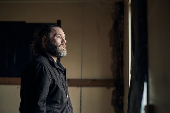 Man met baard kijkt naar buiten door een raam van een vervallen kamer.