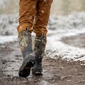 Persoon draagt camouflage Muck Boot laarzen in de sneeuw met een bruine broek.