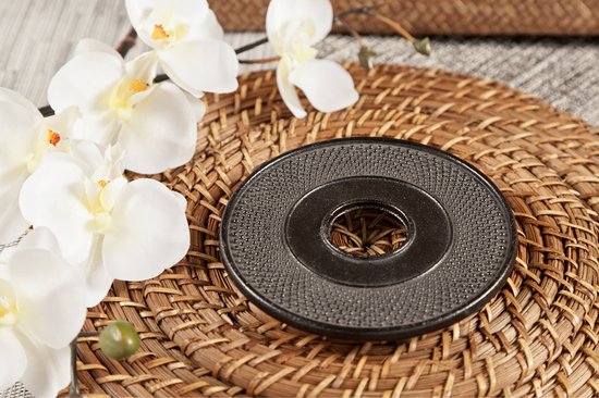 Zwarte gietijzeren onderzetter met puntig patroon op een rieten mat, naast witte orchideeën.