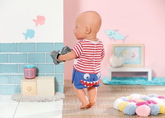 Baby Born pop met rood-wit gestreept shirt met haai en blauwe broek met haaientanden. Houdt grijze haaienschoentjes vast.