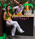 Vrouw in neon-outfit met cocktailketting bij bar, man serveert drankjes bij een tropisch themafeest.