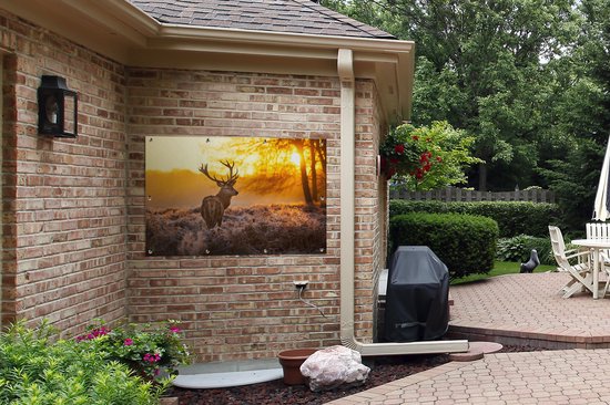 Een tuinposter met een hert in de ochtendzon hangt aan een bakstenen muur buiten.