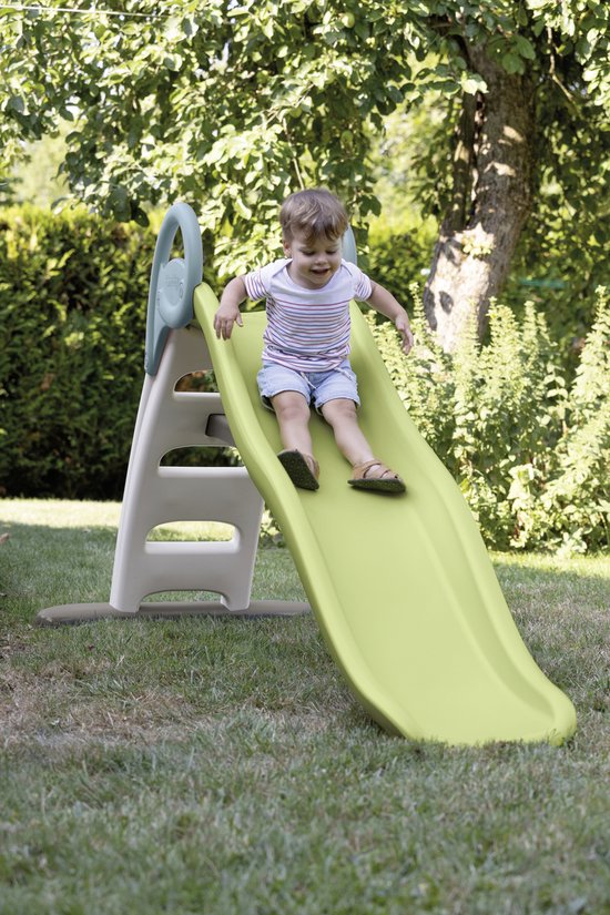 Een jongen glijdt van een groene glijbaan met beige zijkanten in een tuin.