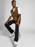Man zittend op een krukje in een bruin gebreid vest, wit T-shirt en zwarte jeans.