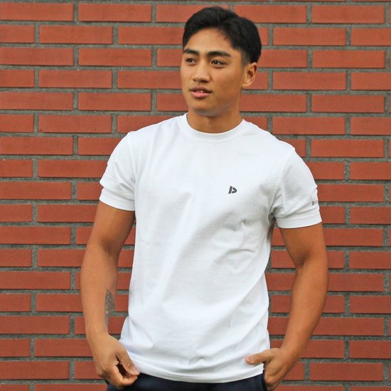 Man met witte Donnay t-shirt en donkerblauwe broek, handen in de zakken, voor een rode bakstenen muur.