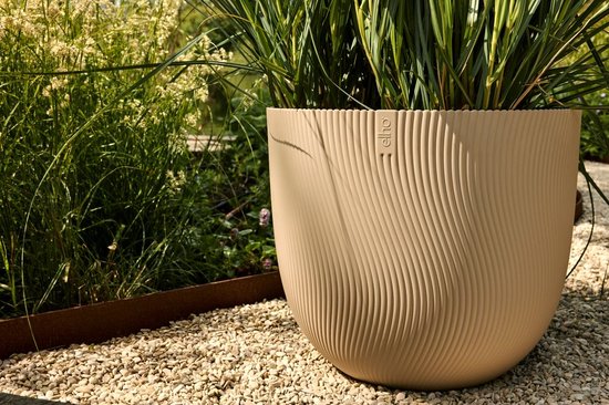 Beige bloempot met golvend ribbelpatroon, op een grindpad naast siergras.