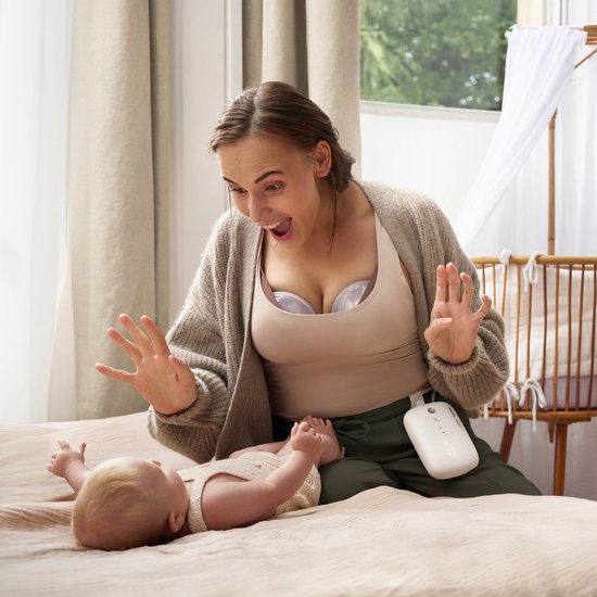 Femme joyeuse portant un tire-lait mains libres Philips Avent, regardant son bébé. L'unité moteur est attachée à son pantalon.