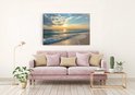 Canvas schilderij met zonsondergang aan zee, hangend boven een roze bank.