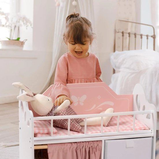 Een jong meisje in een roze jurk speelt met een konijnenknuffel in een wit poppenbedje met roze ruitjes en vlinderdecoraties.