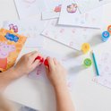 Een kind gebruikt rode Peppa Pig stempels om op een vel papier te drukken, met potloden en andere stempels op de tafel.