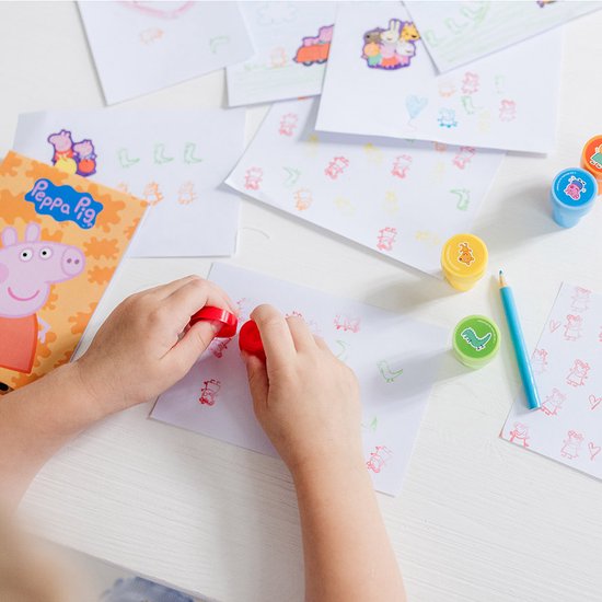 Een kind gebruikt rode Peppa Pig stempels om op een vel papier te drukken, met potloden en andere stempels op de tafel.