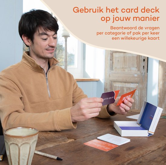 Een man zit aan een tafel en houdt twee kaarten vast. Een open doos met kaarten staat voor hem.