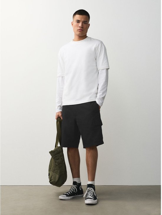 Man met witte longsleeve en zwarte cargoshorts, houdt groene tas vast. Zwarte gympen met witte sokken.