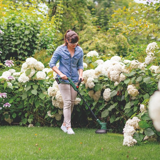 Vrouw met spijkerblouse trimt gras in een tuin met grote witte hortensia's met de Bosch grastrimmer.