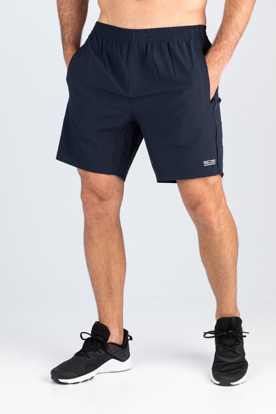 Man in een donkerblauwe Sjeng Sport tennisbroek met beide handen in de zakken en zwarte sportschoenen.