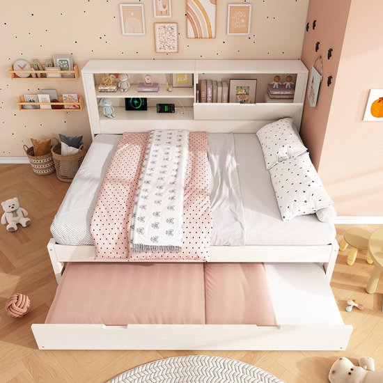 Wit bed met uitschuifbed uitgeschoven. Met roze en witte dekens, en hoofdeinde met opbergvakken.