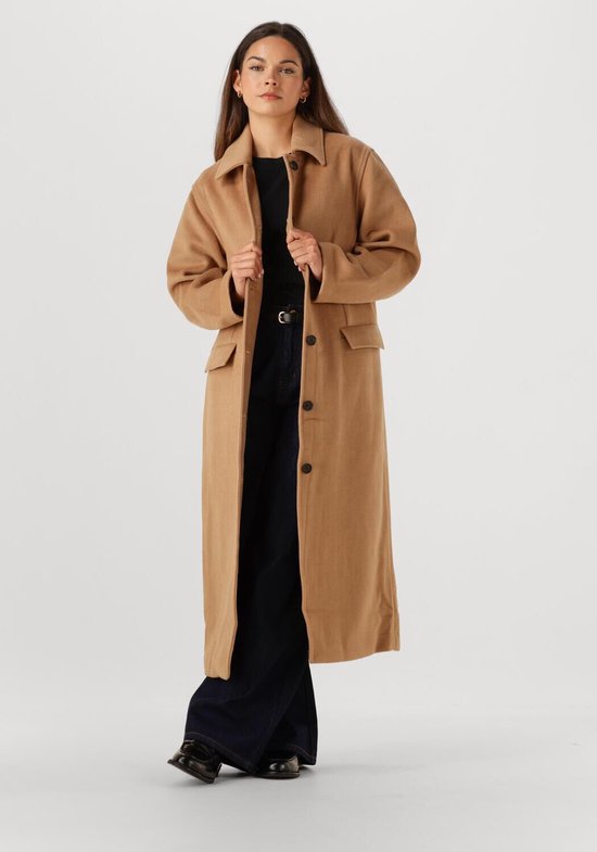 Vrouw draagt een lange beige jas met reverskraag, knoopsluiting en opgestikte zakken. Ze draagt donkere jeans.