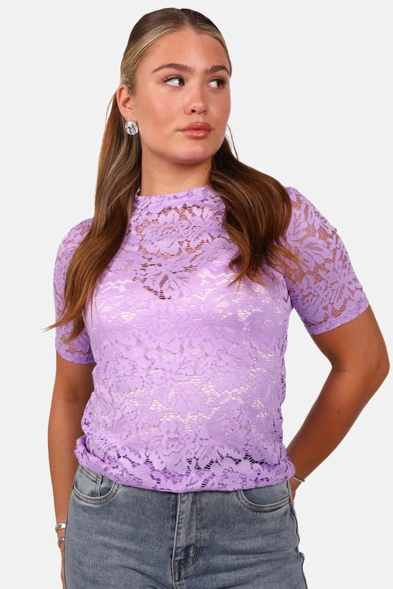 Vrouw met lang bruin haar in lila kanten top met bloemenstructuur, korte mouw, en grijze jeans.