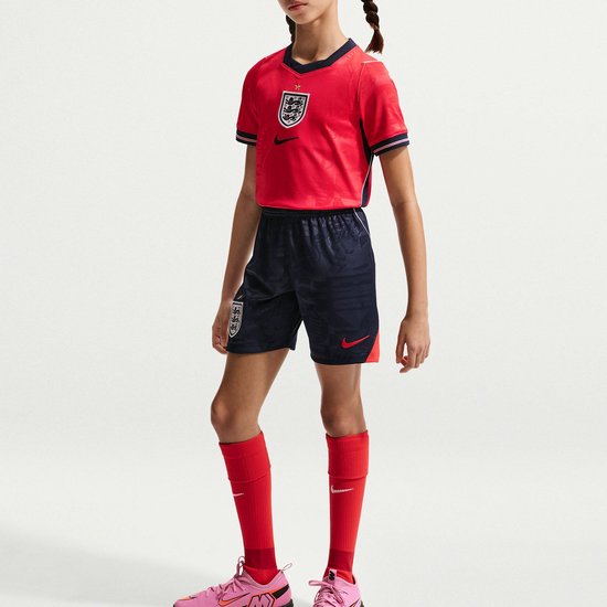 Een meisje in een rood voetbalshirt met marineblauwe shorts en rode sokken. Het shirt heeft een geborduurd wapen en een Nike logo.