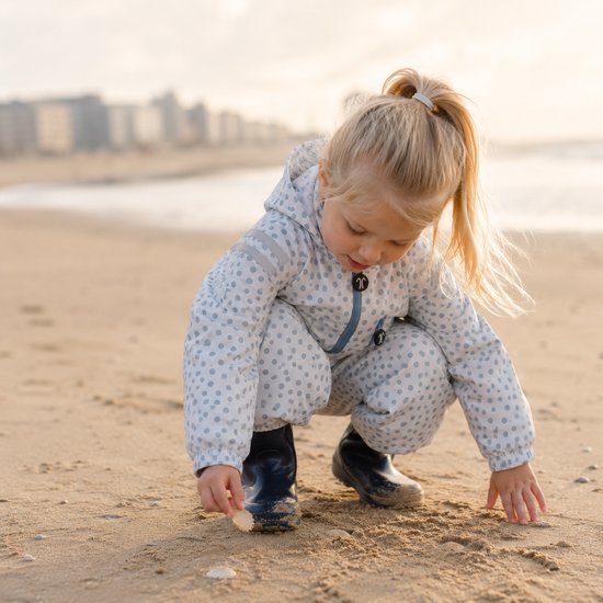Meisje met staartje, hurkend op het strand, in een wit regenpak met lichtblauwe stippen.