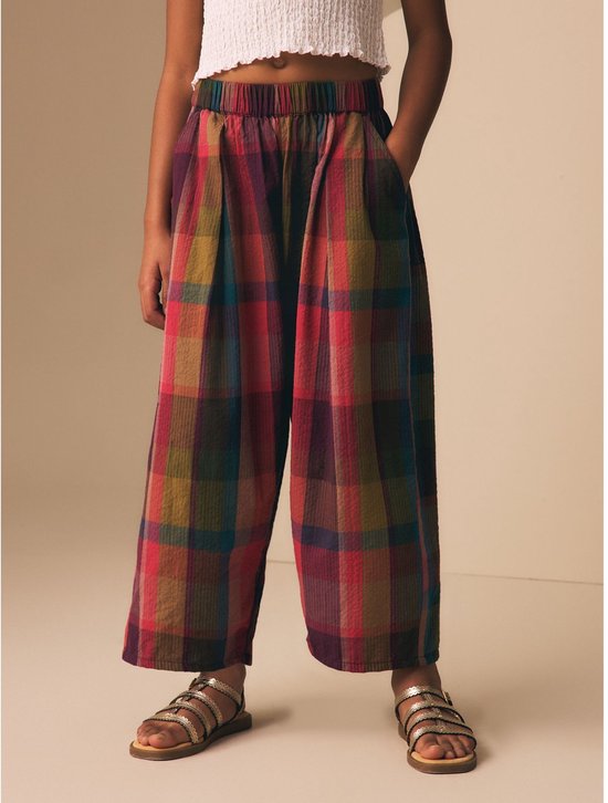 Kind in een geruite broek met wijde, ronde pijpen in rood, paars, groen en bruin.