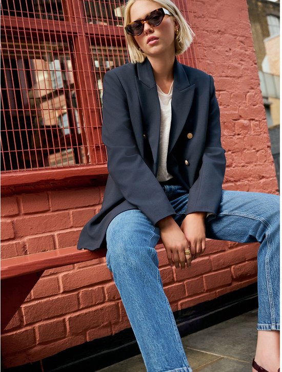 Een vrouw met blond haar, zonnebril, donkerblauw blazer, witte top en spijkerbroek, zittend op een rode bank.