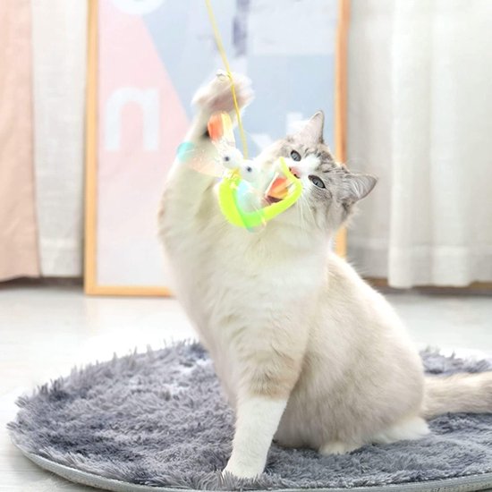 Een witte en grijze kat met blauwe ogen vangt speels een hangend, felgekleurd libel-speeltje met grote, ronde ogen.