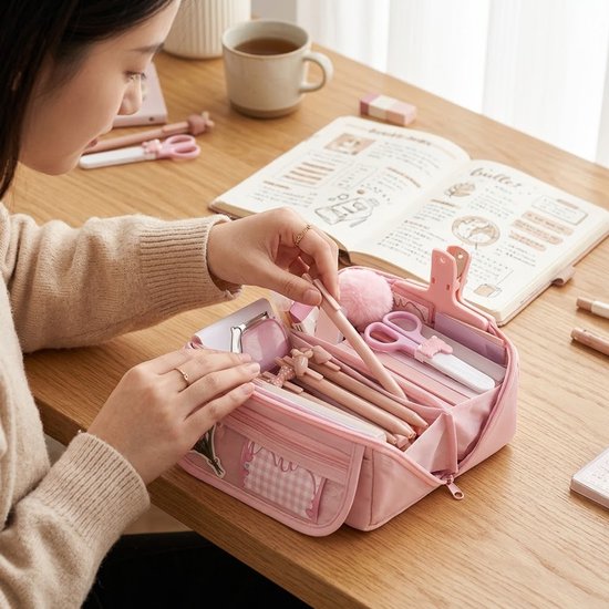 Roze etui met dubbele rits, gevuld met pennen, schaar en andere kantoorspullen. Een vrouw pakt een pen uit de etui. Voorvak met ruitjes en bloemdecoratie.