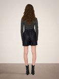 Vrouw met lang bruin golvend haar, draagt een gestreepte grijze coltrui en zwarte faux leren shorts.
