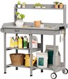 Grijze houten werktafel met planten, potten, flessen, mand, gieter en tuingereedschap. Twee zakken met groene labels staan op de onderste plank.