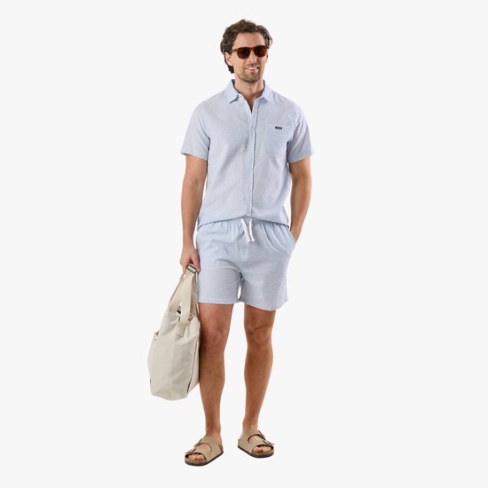 Man in lichtblauwe gestreepte seersucker pyjamaset met korte broek en korte mouwen, met beige sandalen en een witte tas.