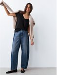 Model met een beige vest over een zwart T-shirt met V-hals en een wijde jeans.