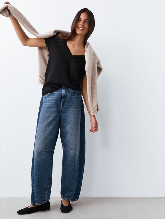 Model met een beige vest over een zwart T-shirt met V-hals en een wijde jeans.