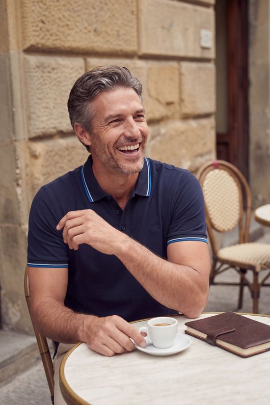 Man geniet van koffie in een marineblauwe poloshirt met geborduurd logo en blauw-witte strepen.