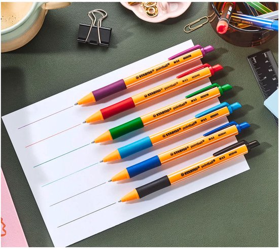 Zes STABILO pointball pennen met oranje hulzen en gekleurde handvatten, liggend op wit papier met gekleurde lijnen.
