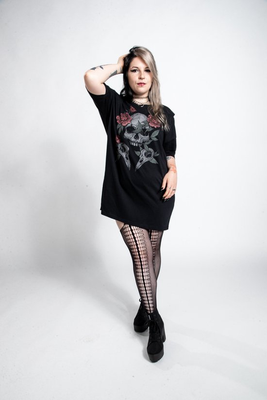 Vrouw met grijze haren, tatoeages, visnetkousen en zwarte laarzen draagt een zwart oversized T-shirt met schedel- en rozenprint.