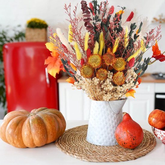 Gedroogd herfstboeket in een metalen kan op een rieten onderzetter met pompoenen en een rode koelkast.