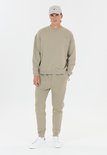 Man in beige trainingsbroek en sweater met logo, met een beige pet.