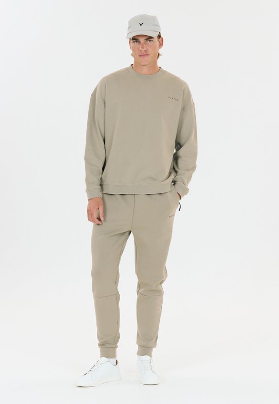 Man in beige trainingsbroek en sweater met logo, met een beige pet.