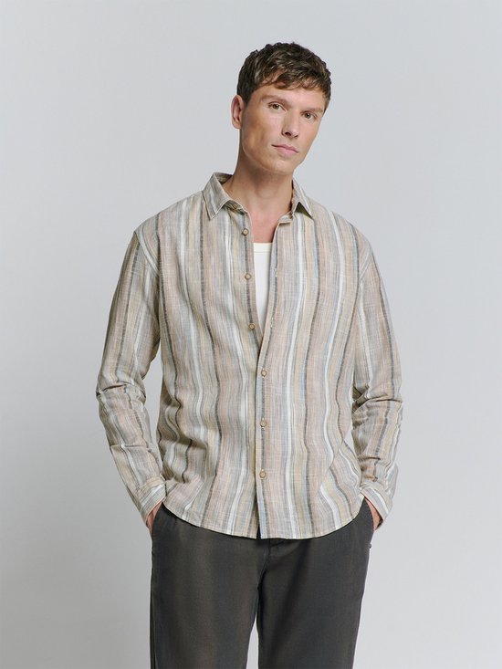 Model in een langere, loszittende beige linnen mix blouse met verticale strepen in diverse kleuren, waaronder bruin, blauw en geel. Een witte tanktop is zichtbaar onder de blouse.