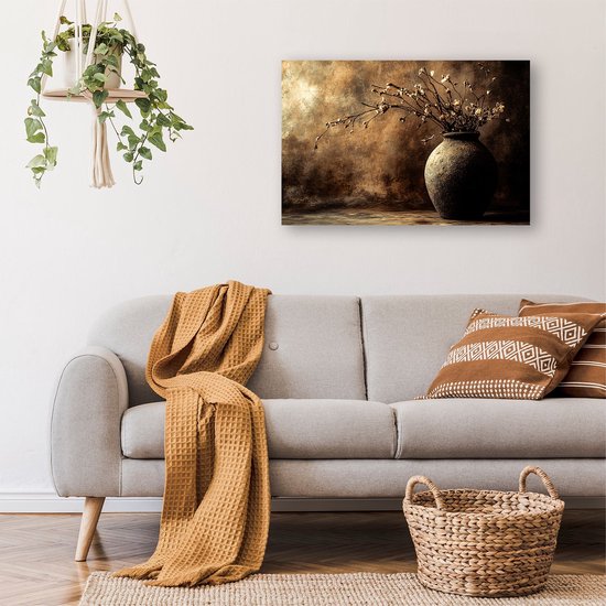 Canvasafdruk met een donkere, aardse vaas gevuld met gedroogde bloemen. Hangt boven een grijze bank met een mosterdgele deken en kussens.
