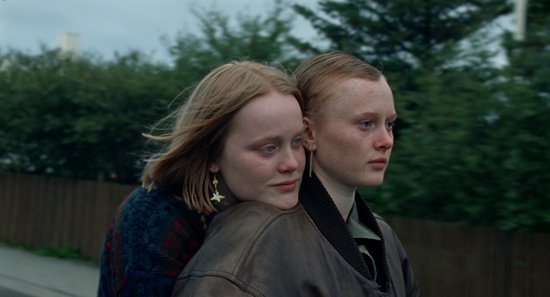 Twee jongedames met sproeten kijken omhoog. De ene met roodachtig haar, de andere met kort blond haar. Buiten, groenige achtergrond.