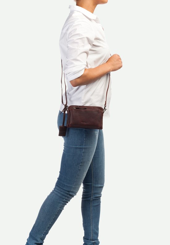 Bruine leren schoudertas van Burkely met een lange donkerbruine band, gedragen door een model in een wit shirt en spijkerbroek.