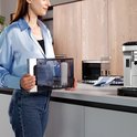 Vrouw met blauwe blouse houdt een transparante watertank met wit waterfilter vast naast een koffiezetapparaat.