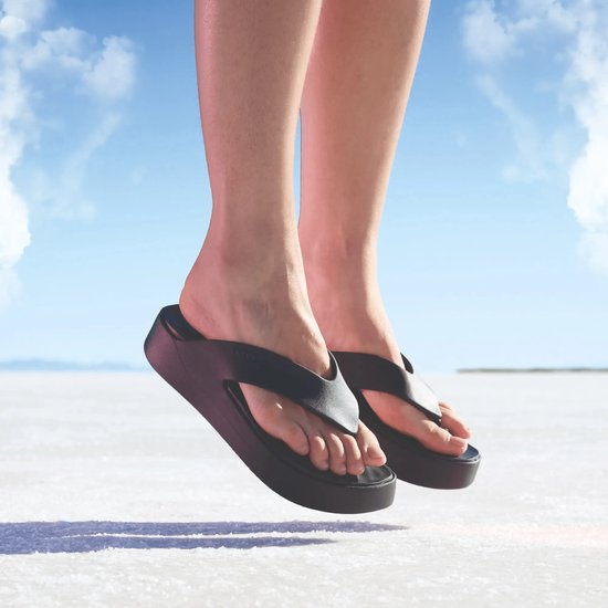 Pieds portant des tongs compensées noires Crocs flottant au-dessus d'une surface blanche type désert de sel.