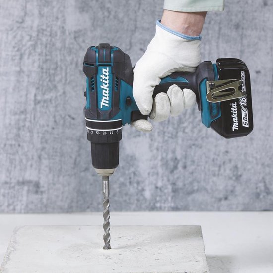 Hand met witte handschoenen gebruikt een turquoise en zwarte Makita accuboormachine om in beton te boren.