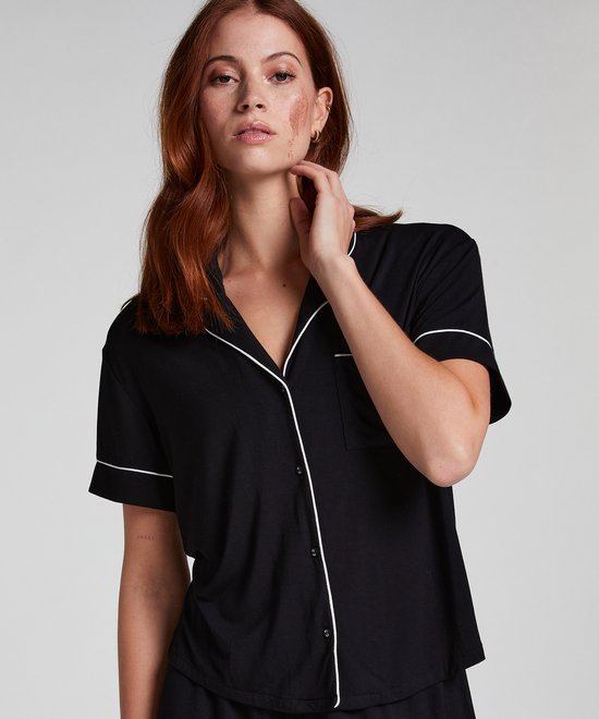 Model met rood haar draagt een zwart pyjamashirt met korte mouwen en witte piping. Het shirt heeft een open kraag en borstzakje.