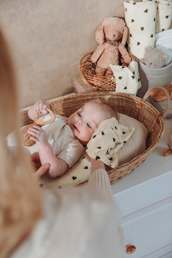 Baby met wit rompertje in rieten mand, speelt met rammelaar. Handdoekje met zwarte hartjes.