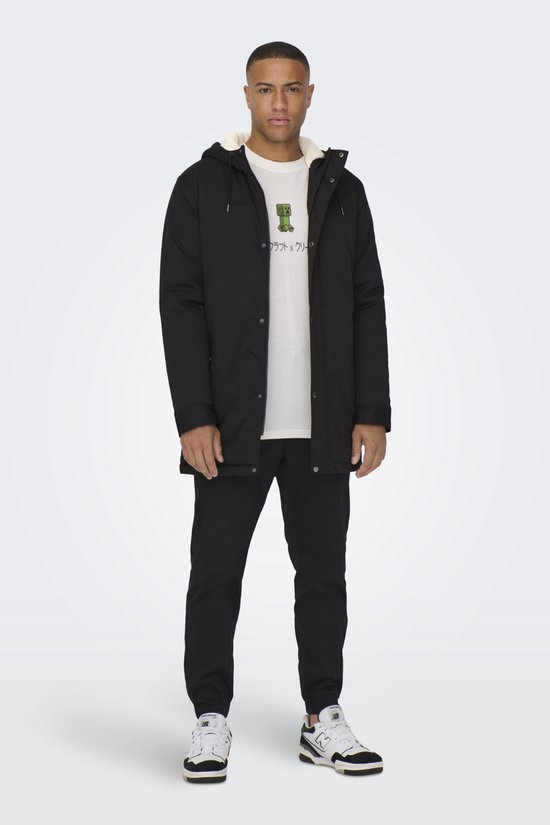 Staande man in zwarte parka met witte voering, wit Minecraft T-shirt, zwarte broek en sneakers.